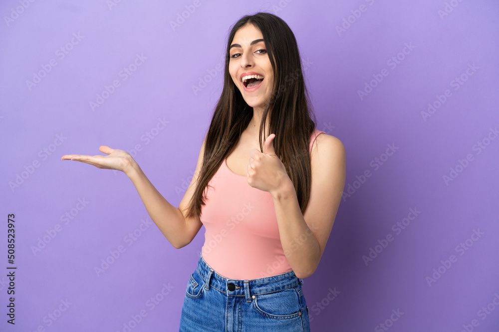 Young caucasian woman isolated on purple background holding copyspace imaginary on the palm to insert an ad and with thumbs up