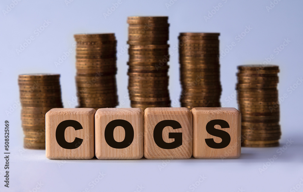 COGS - acronym on wooden cubes on the background of stacks of coins