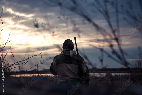 Hunter man in camouflage with shotgun looking into the distance with flying gooses on horizon during dramatic sunset during hunting season 