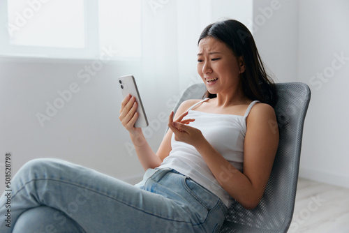 Irritated angry cute young Asian woman quarrel with boyfriend in messages sit on chair at home interior living room. Distance communication, Remote work, Social media concept. Cool offer Banner Mockup