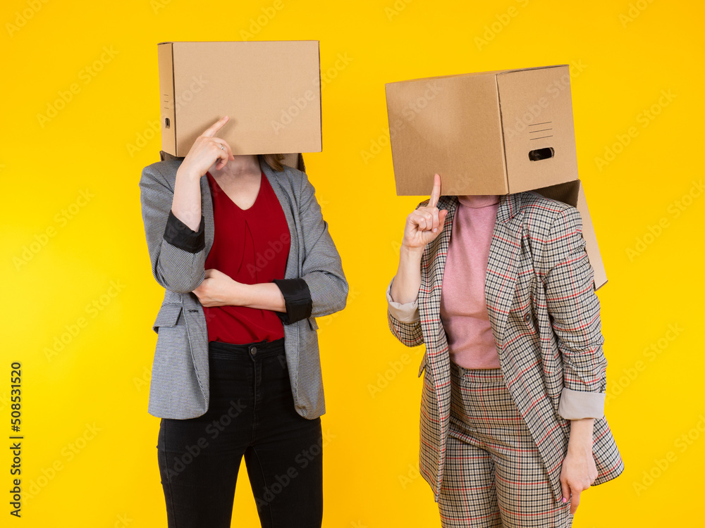 Women gossip. Two girls with cardboard boxes on their faces. Women hide  their heads in boxes. Concept of gossip between girls. Gossip girl on  yellow background. Female dressed in business style Stock