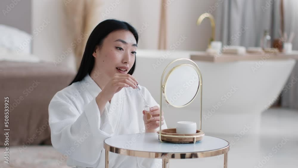 Skin care and vitamization. Young pretty asian lady applying nourishing serum on face looking at mirror, free space Skin care and vitamization. Young pretty asian lady applying nourishing serum on face looking at mirror, free space