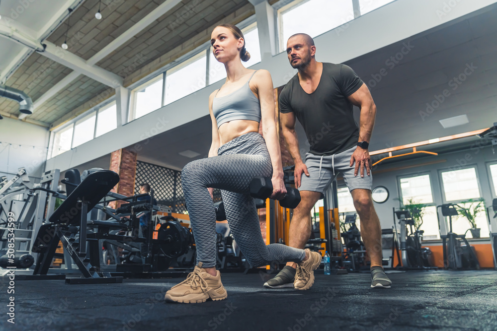 Gym time. Horizontal indoor wide shot of two caucasian people. Attractive pretty young woman doing exercise with weigths, good-looking hairless middle-aged man standing behind her. High quality photo