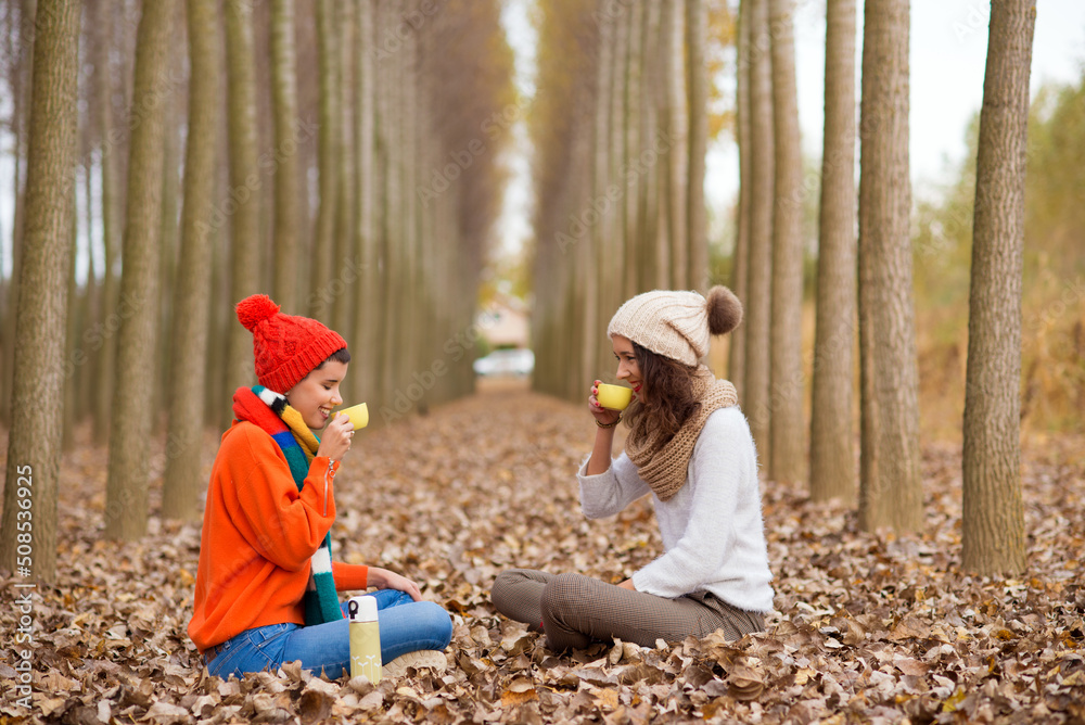 Two smiling girls drinking coffee while sitting on the forest autumn leaves on a beautiful day Two smiling girls drinking coffee while sitting on the forest autumn leaves on a beautiful day