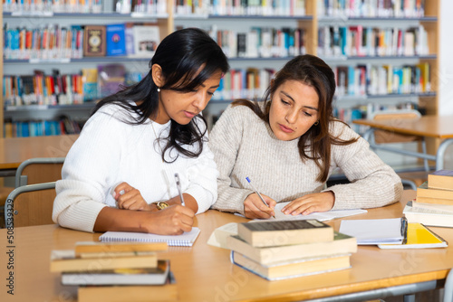 Two young adult women gaining new skills at public library, reading books and making notes