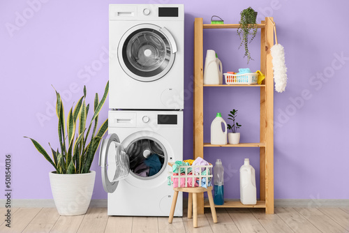 Modern drying, washing machines, basket with laundry and shelf unit near violet wall © Pixel-Shot