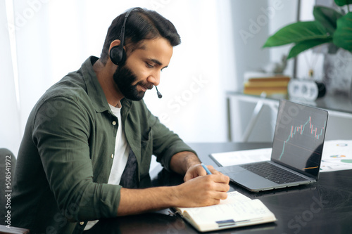 Online trading and stock market concept. Indian or arabian man focused analyze stocks price on online marketplace before buying, uses laptop, while sitting in his modern office