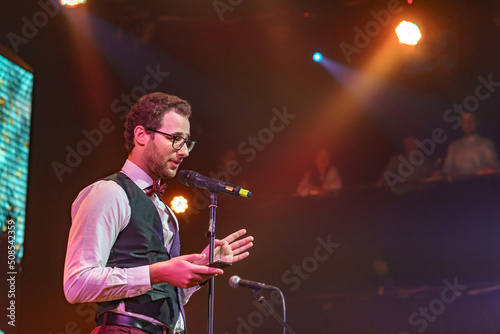 Attractive and confident Public speaker with glasses bow tiea a mobile phone in hand in the spotlight talking in conference or concert hall. People blurred in background