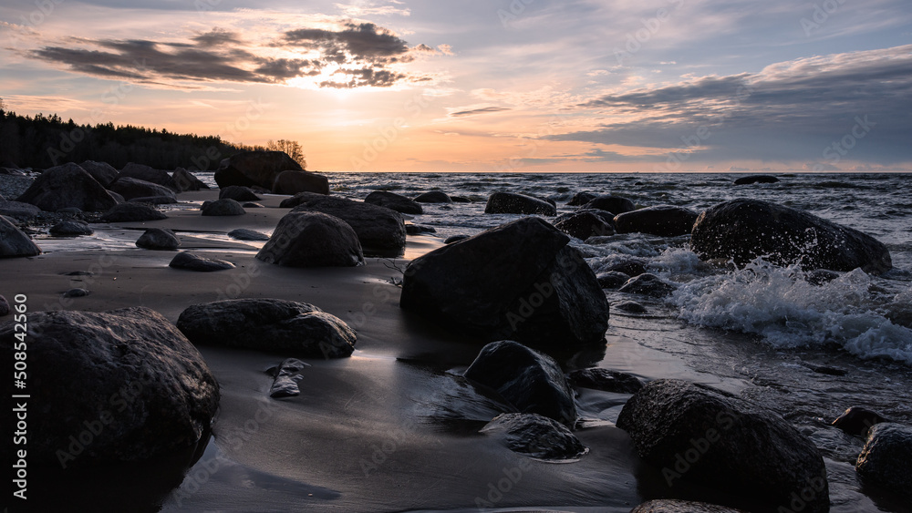 Seashore at sunset. Granite boulders on a sandy beach are washed by restless waves, backlight.