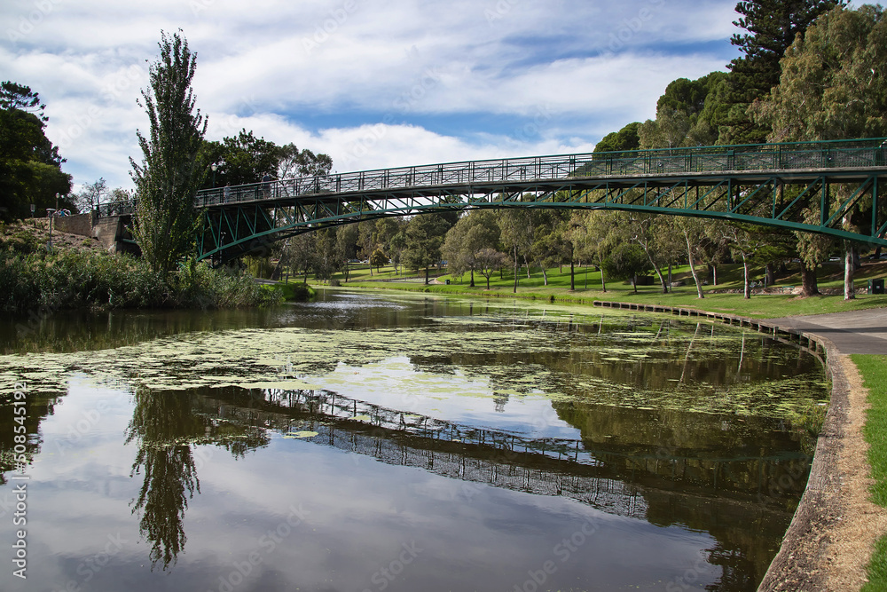 Fototapeta premium Bridge over the river in the city park surrounded by green trees with reflection in the water, cityscape