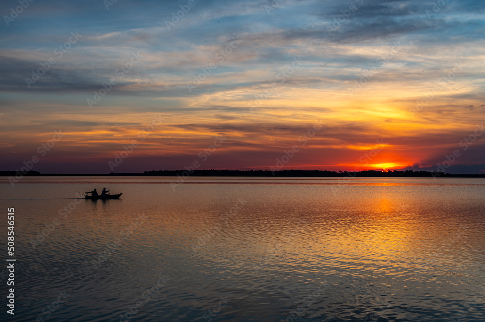 Sun Setting over Lake with Rippled Water, Colorful Sky, and Boat Silhouette