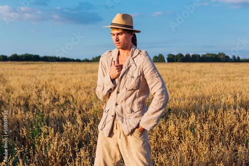 Tall handsome man dressed in a coarse linen suit and hat standing at golden oat field © Smile