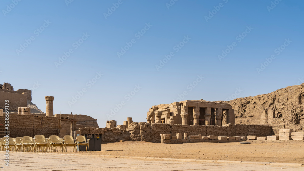 A fragment of the ancient temple of Horus in Edfu. Dilapidated walls and colonnades are visible. Chairs for the audience stand in a row on the square. Clear blue sky. Copy space. Egypt