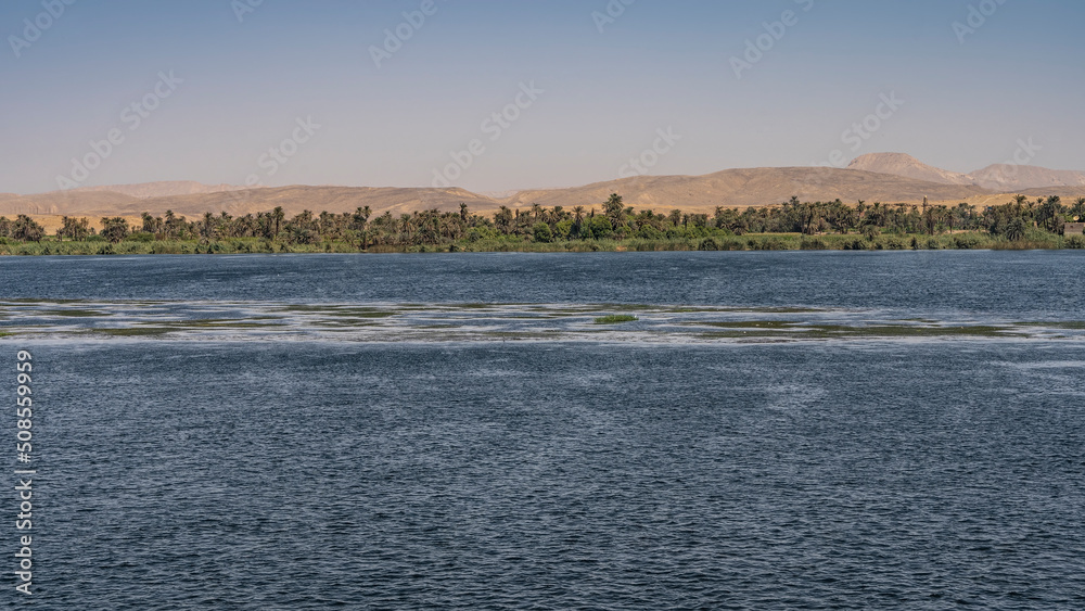 The wide and calm Nile River. Ripples on the blue water. There is green vegetation on the shore. Sand dunes against the sky. Egypt