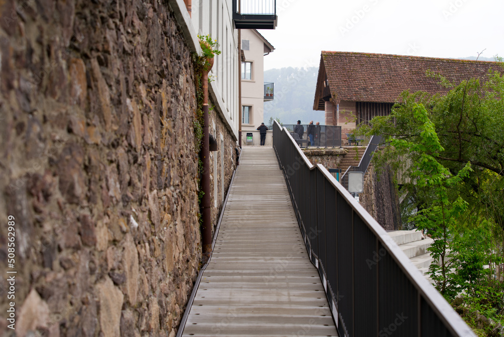Pathway along a stone wall to wooden covered bridge at Village of Stein, Canton Aargau, on a cloudy spring day. Photo taken May 6th, 2022, Stein, Switzerland.