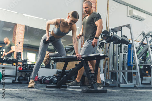 Fototapeta Naklejka Na Ścianę i Meble -  An attractive caucasian young adult woman using free weights at a fitness facility to build up her upper body strength under the guidance of her male private personal trainer. High quality photo