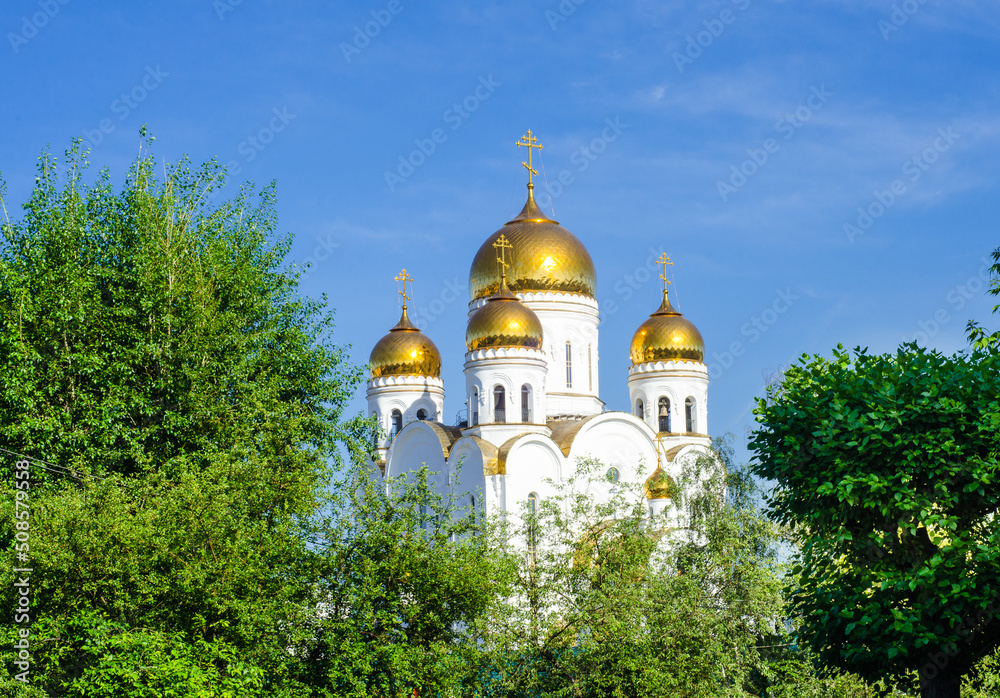 golden domes of  Orthodox Church against blue sky surrounded by green trees.