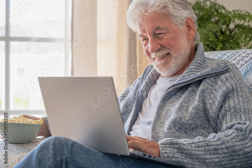 Elderly 70s man seated on sofa browsing on laptop, senior looking at laptop screen surfing the net, older generation and modern tech application easy usage concept