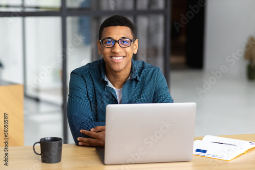 Portrait of successful African American man entrepreneur with glasses working using laptop computer sitting in modern office looking at the camera and smiles friendly