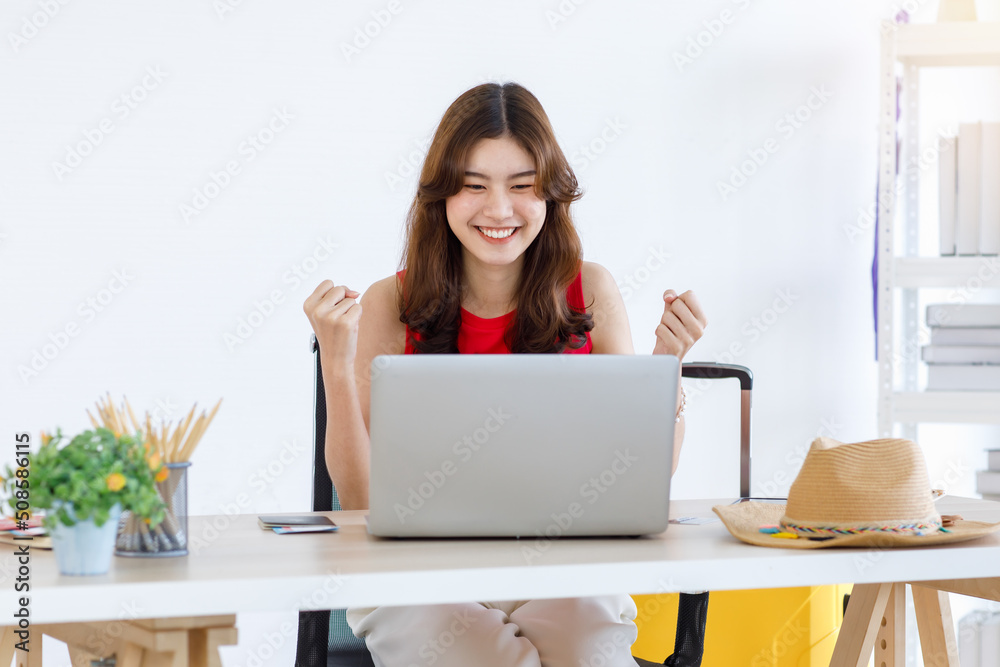 Asian excited happy female traveler in casual outfit sitting looking at laptop computer holding fists up celebrating after lucky winning free travelling trip vacation holiday package prize online Asian excited happy female traveler in casual outfit sitting looking at laptop computer holding fists up celebrating after lucky winning free travelling trip vacation holiday package prize online