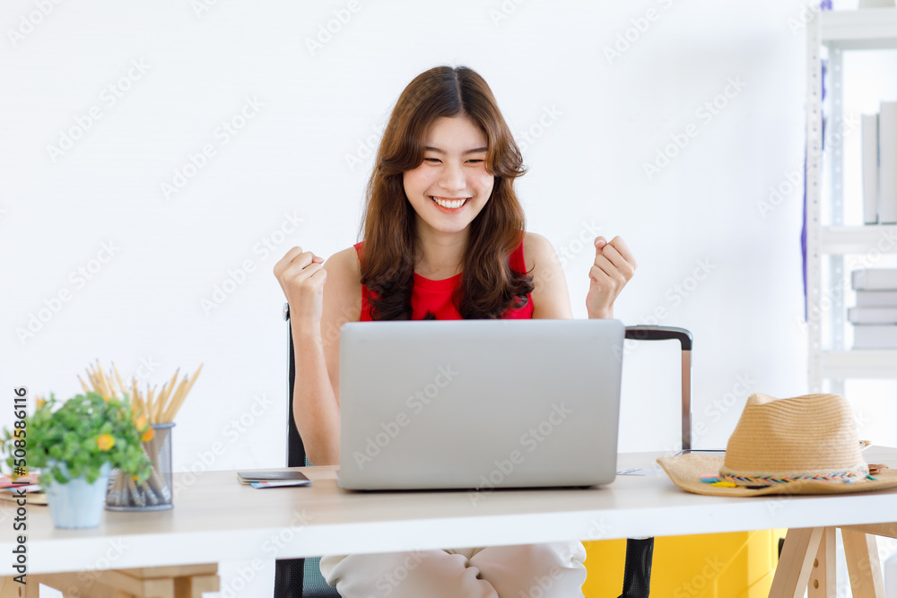 Asian excited happy female traveler in casual outfit sitting looking at laptop computer holding fists up celebrating after lucky winning free travelling trip vacation holiday package prize online Asian excited happy female traveler in casual outfit sitting looking at laptop computer holding fists up celebrating after lucky winning free travelling trip vacation holiday package prize online