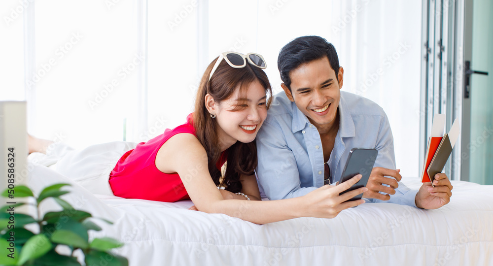Asian young traveler lover couple in casual outfit with sunglasses and hat laying down smiling together on bed holding passport boarding pass ticket using smartphone booking hotel reservation online Asian young traveler lover couple in casual outfit with sunglasses and hat laying down smiling together on bed holding passport boarding pass ticket using smartphone booking hotel reservation online