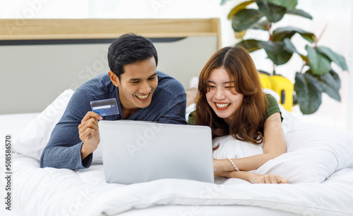 Asian happy romantic male female husband and wife lover couple lay down smiling together on pillow under blanket on bed using laptop notebook computer and credit card shopping making payment online © Bangkok Click Studio