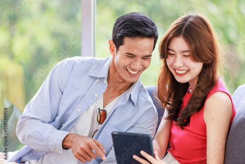 Asian young happy lovely boyfriend and girlfriend couple in casual outfit sitting smiling on cozy sofa couch in living room using tablet computer watching streaming movie online together in weekend