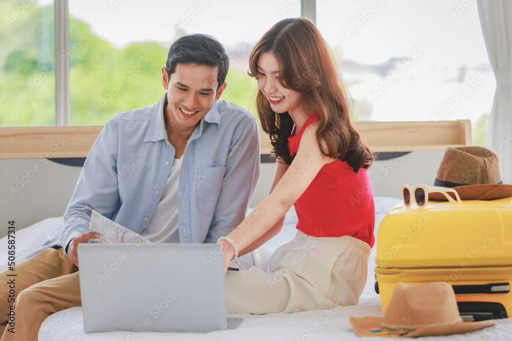 Asian young romantic lover couple boyfriend and girlfriend in casual outfit sitting smiling helping packing luggage together on bed while male using tablet computer booking vacation trip hotel online