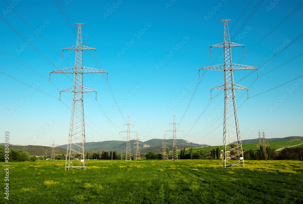 High voltage lines and power pylons in a flat and green agricultural mountain landscape on a sunny day with blue sky.