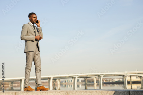 Serious pensive young black businessman with beard concentrated on phone conversation walking over city riverside