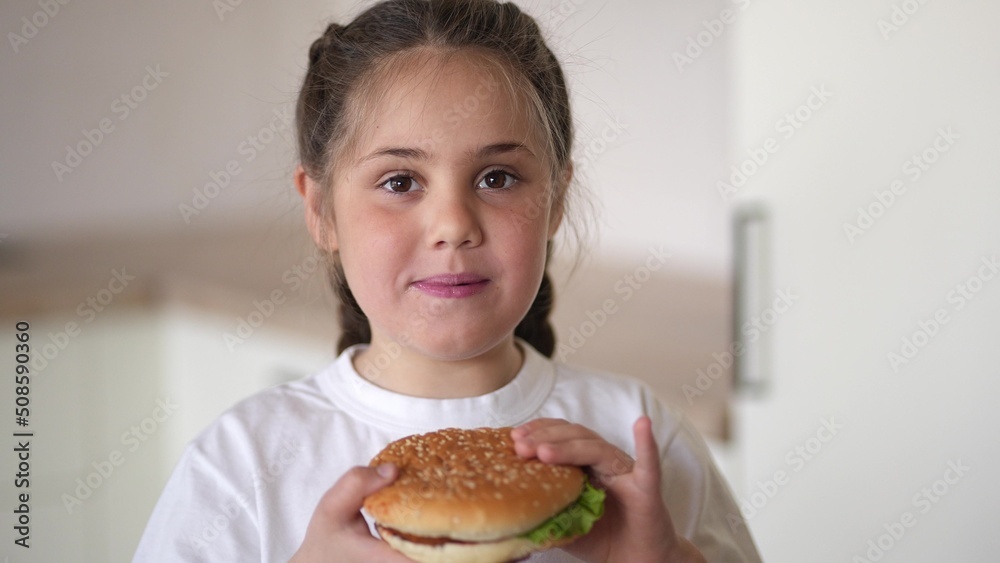 little girl eating a hamburger. unhealthy fast food proper nutrition concept. child greedily with pleasure bites a big burger in the kitchen at home. lifestyle kid eats fast food close-up little girl eating a hamburger. unhealthy fast food proper nutrition concept. child greedily with pleasure bites a big burger in the kitchen at home. lifestyle kid eats fast food close-up
