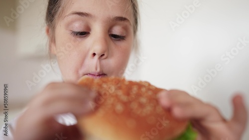 little girl eating a hamburger. unhealthy fast food proper nutrition concept. child greedily with pleasure bites a big burger in the kitchen at home. kid eats fast food meal close-up