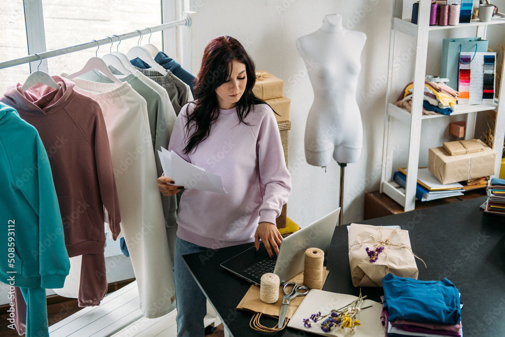 Young woman tailor fashion designer dressmaker using laptop in seamstress studio Young woman tailor fashion designer dressmaker using laptop in seamstress studio