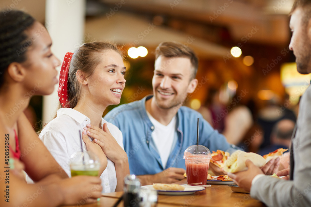 Attractive girl with ribbon in hair sitting at table in cafe and pointing at herself while introducing herself to new friends