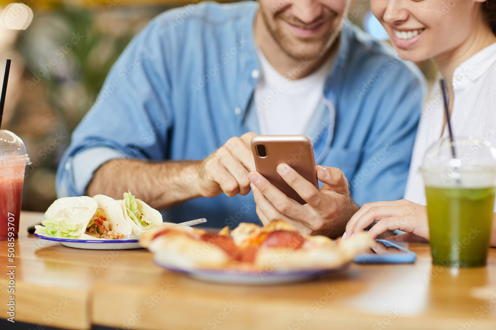 Close-up of positive young couple sitting at table with dishes and surfing social media on phone Close-up of positive young couple sitting at table with dishes and surfing social media on phone
