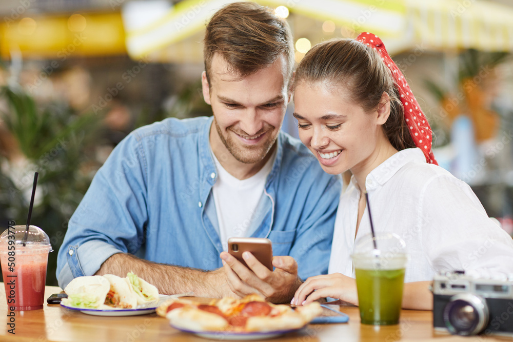 Jolly young couple in casual clothing sitting at table and using smartphone while watching video on social media