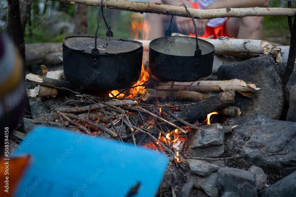 A campfire with two black cast iron pots hanging above it, one with boiling water, surrounded by wood and stones, creating a warm atmosphere A campfire with two black cast iron pots hanging above it, one with boiling water, surrounded by wood and stones, creating a warm atmosphere