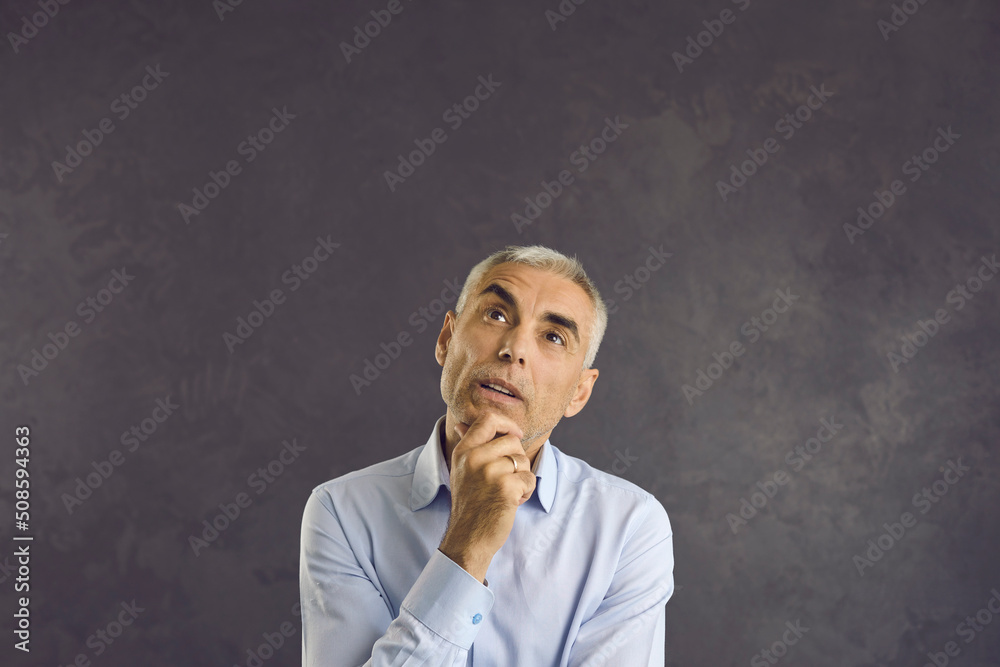 Studio headshot portrait of senior questionable man who is looking up thinking or planning. Thoughtful focused man holding his chin trying to remember something or make choice. Gray background.