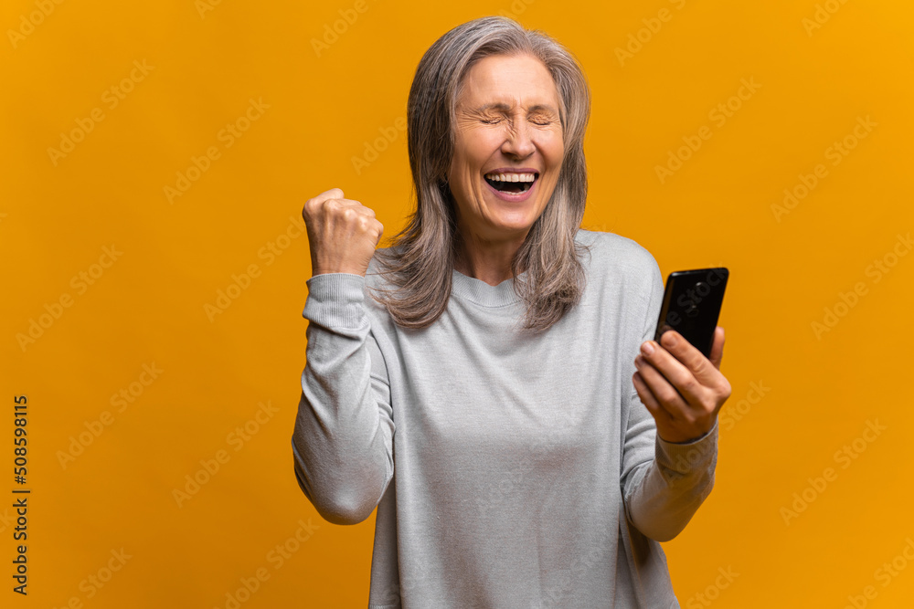 Happy satisfied woman holding smartphone and smiling making yes gesture, celebrating online lottery or giveaway victory. Indoor studio shot isolated on yellow background Happy satisfied woman holding smartphone and smiling making yes gesture, celebrating online lottery or giveaway victory. Indoor studio shot isolated on yellow background