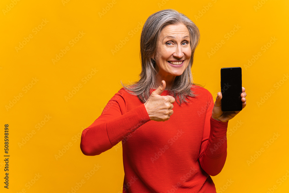 Delighted by the holiday deal mature woman holding smartphone with empty screen. Overjoyed middle-aged lady demonstrates phone with blank display and showing big finger isolated on yellow Delighted by the holiday deal mature woman holding smartphone with empty screen. Overjoyed middle-aged lady demonstrates phone with blank display and showing big finger isolated on yellow