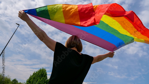 LGBTQIA month pride, rainbow peace flag against blue sky with clouds on a sunny day. Hold woman in hand and Celebrate Bisexuality Day or National Coming Out Day