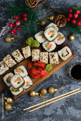 Large set of sushi on a wooden board top view. Rolls with sesame seeds, nori, cheese, red fish, cucumber and eel, ginger, wasabi, soy sauce, bamboo sticks and christmas decor on gray background