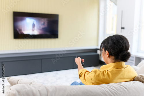 Happy young multiracial woman watching television programs, sits on comfortable couch with remote controller, switching channels, looks at the screen, girl spends leisure at home