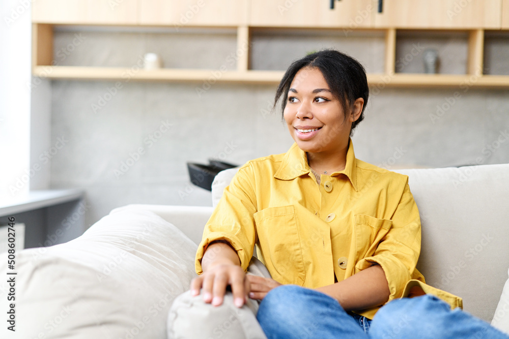 Carefree dark skin lady looks away dreamily, rest, enjoying calm at home. Serene African-American young woman leaned back on the sofa