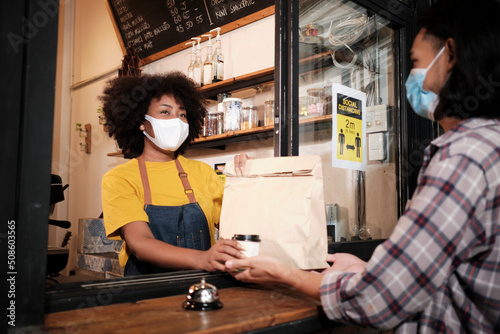 African American female barista with face mask works in the cafe with social distance, takeaway coffee for a customer, new normal service of small business coffee shop in COVID19 quarantine lifestyle.