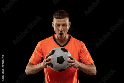 Front camera view of young male soccer player in orange-blue football kit posing with ball isolated on dark background. Concept of sport, goals, competition, emotions