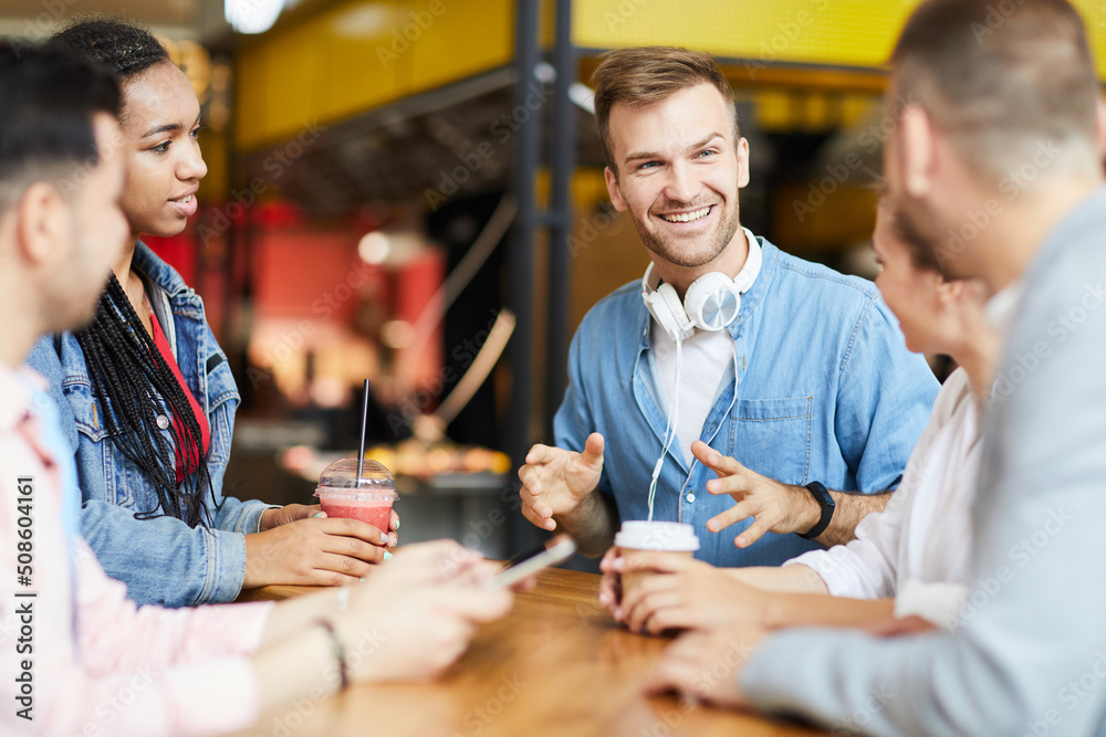 Smiling handsome young man with stubble sitting at table and gesturing hands while spending leisure with friends in cafe