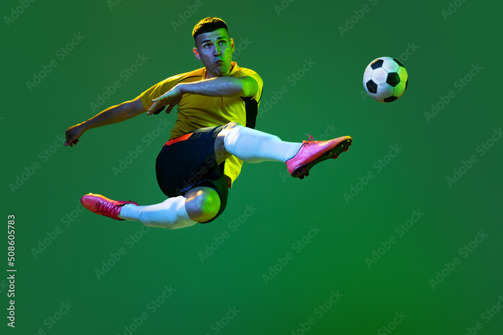 Studio shot of young professional male football soccer player in motion isolated on green background in neon. Concept of sport, goals, competition, hobby, achievements