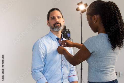 Putting Lavalier Microphone on Caucasian Male Wearing Business Attire. Video Production or Film Set Behind the Scenes. Mic Clips Onto Shirt. Lighting Seen in Background